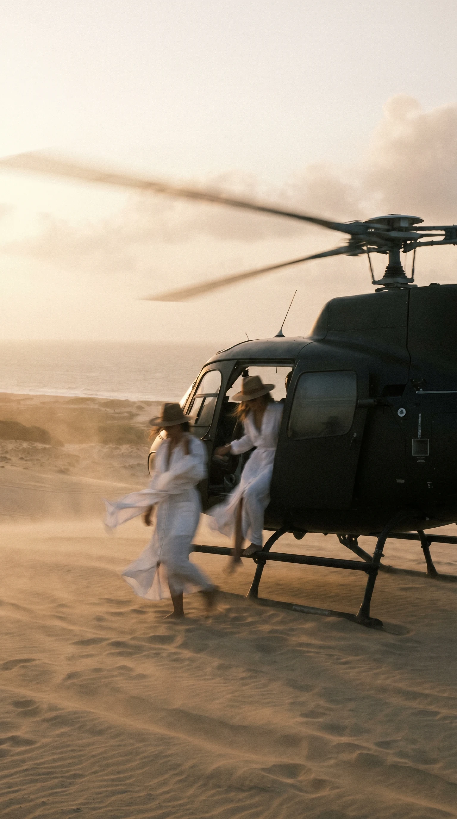 Duas convidadas embarcam em um helicóptero parado na areia, luz dourada do fim de tarde.